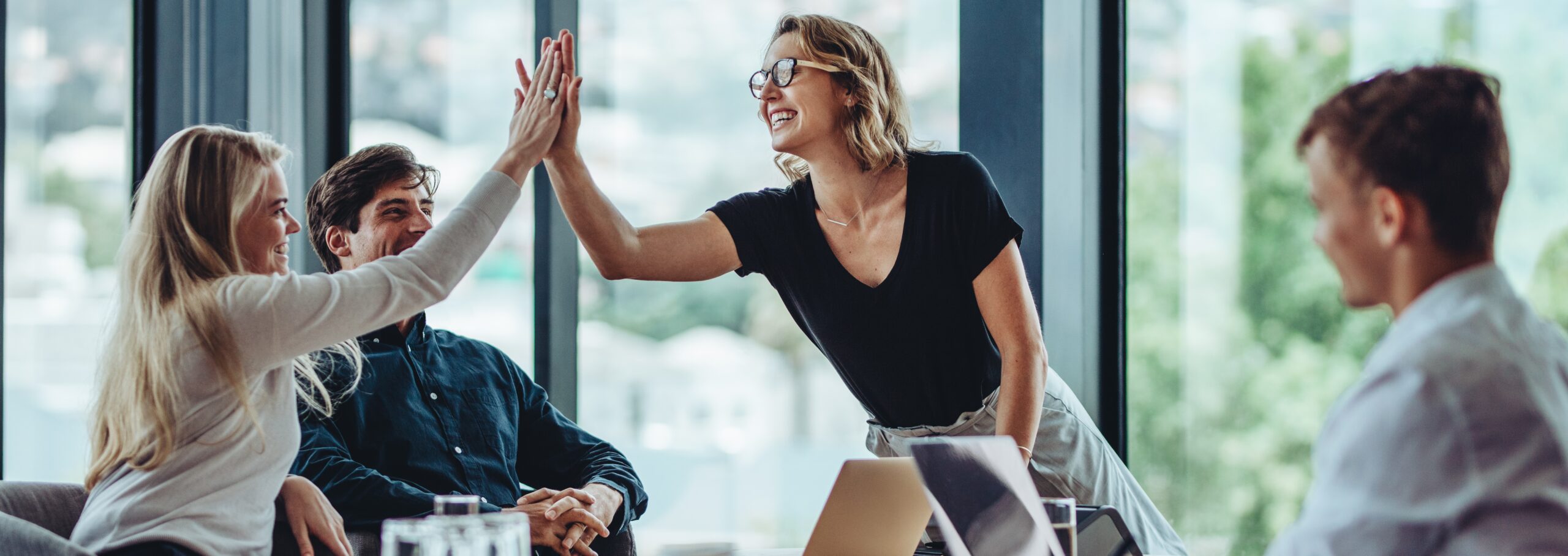 Team feiert gemeinsamen Erfolg im Büro: Zwei Mitarbeiterinnen geben sich ein High-Five, während Kollegen lächeln und zusammenarbeiten